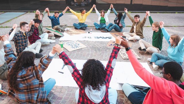A group of multicultural peacefully activists are sitting with posters outside.